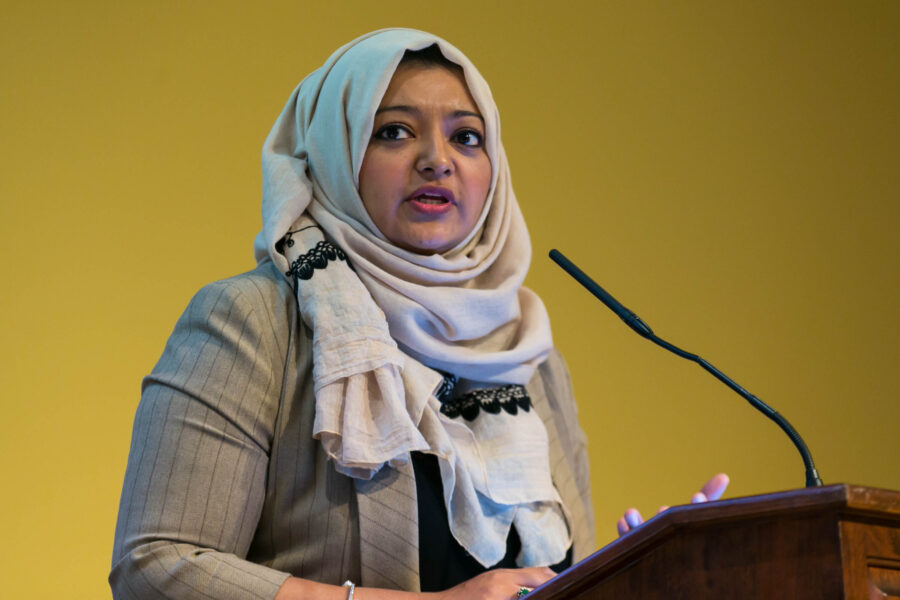 Rabia Chaudry wearing a beight short and head scarf speaks into a microphone.