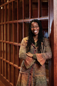 Portrait of Tiya Miles, a Black woman with long black hair, smiling with arms crossed in a library wearing a brown and green knit sweater.